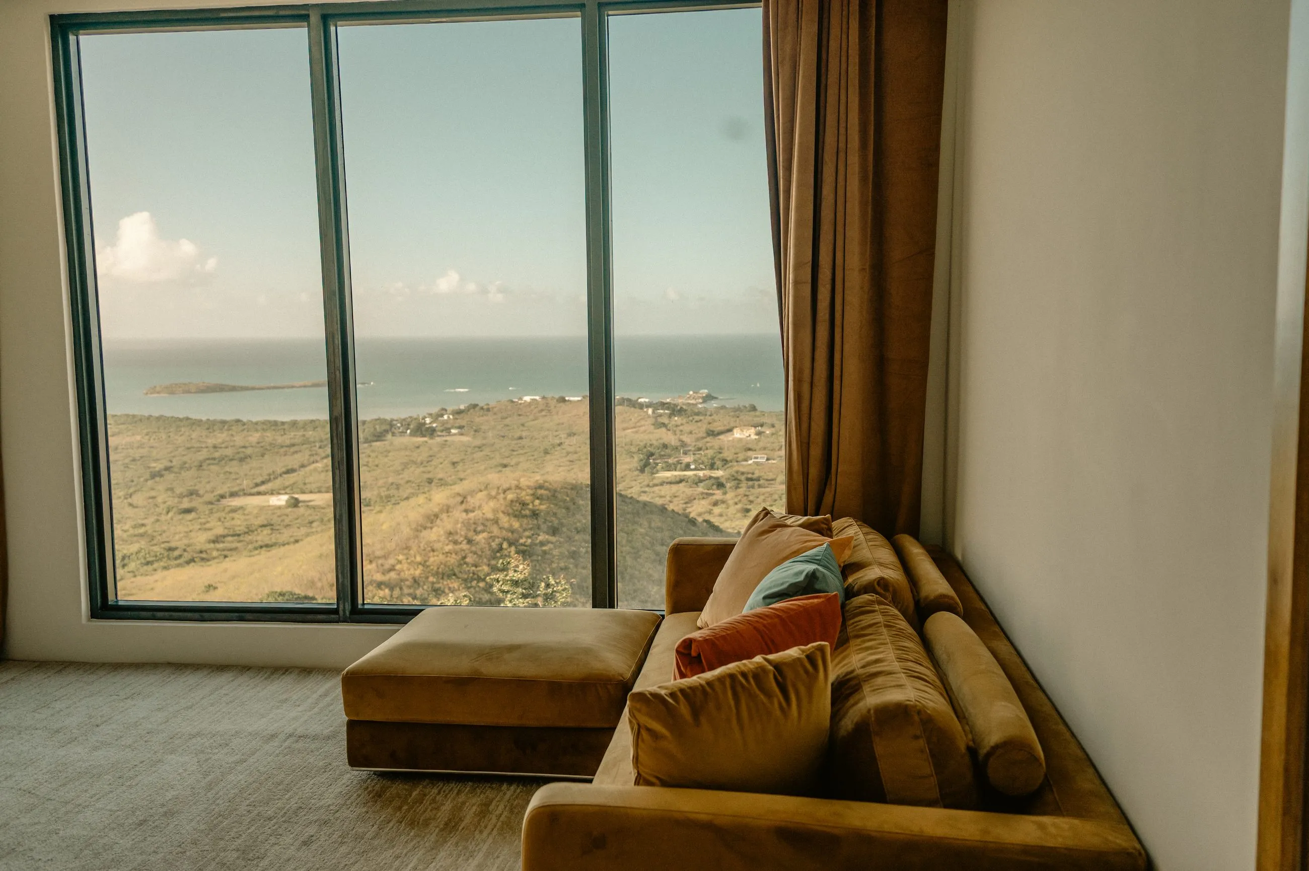 Living area with floor-to-ceiling windows framing the hills and the Caribbean Sea, low sectional sofa and stone-tile floor