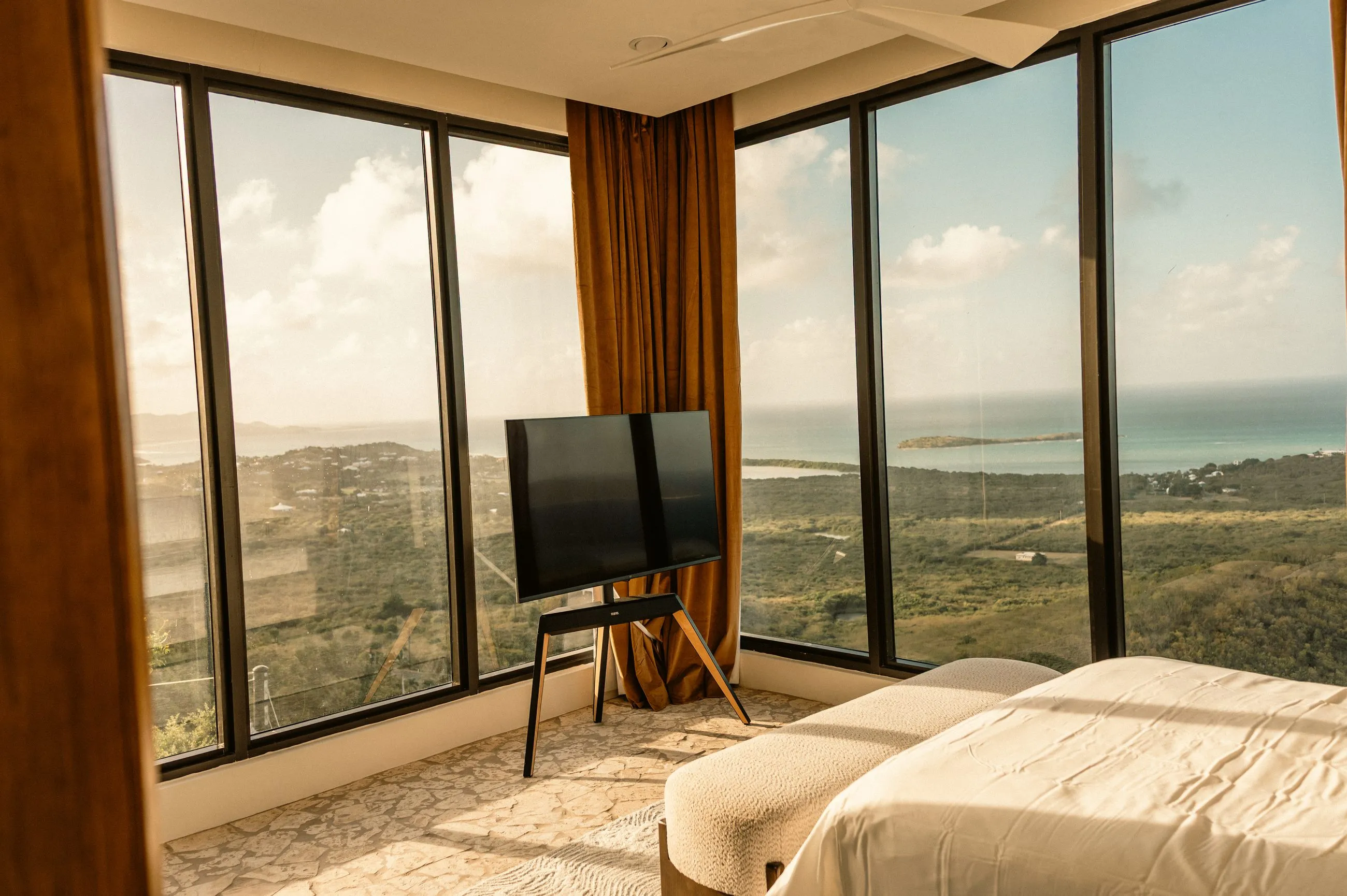 Primary bedroom with corner floor-to-ceiling windows looking out over green hills to the sea, king bed in cream linens