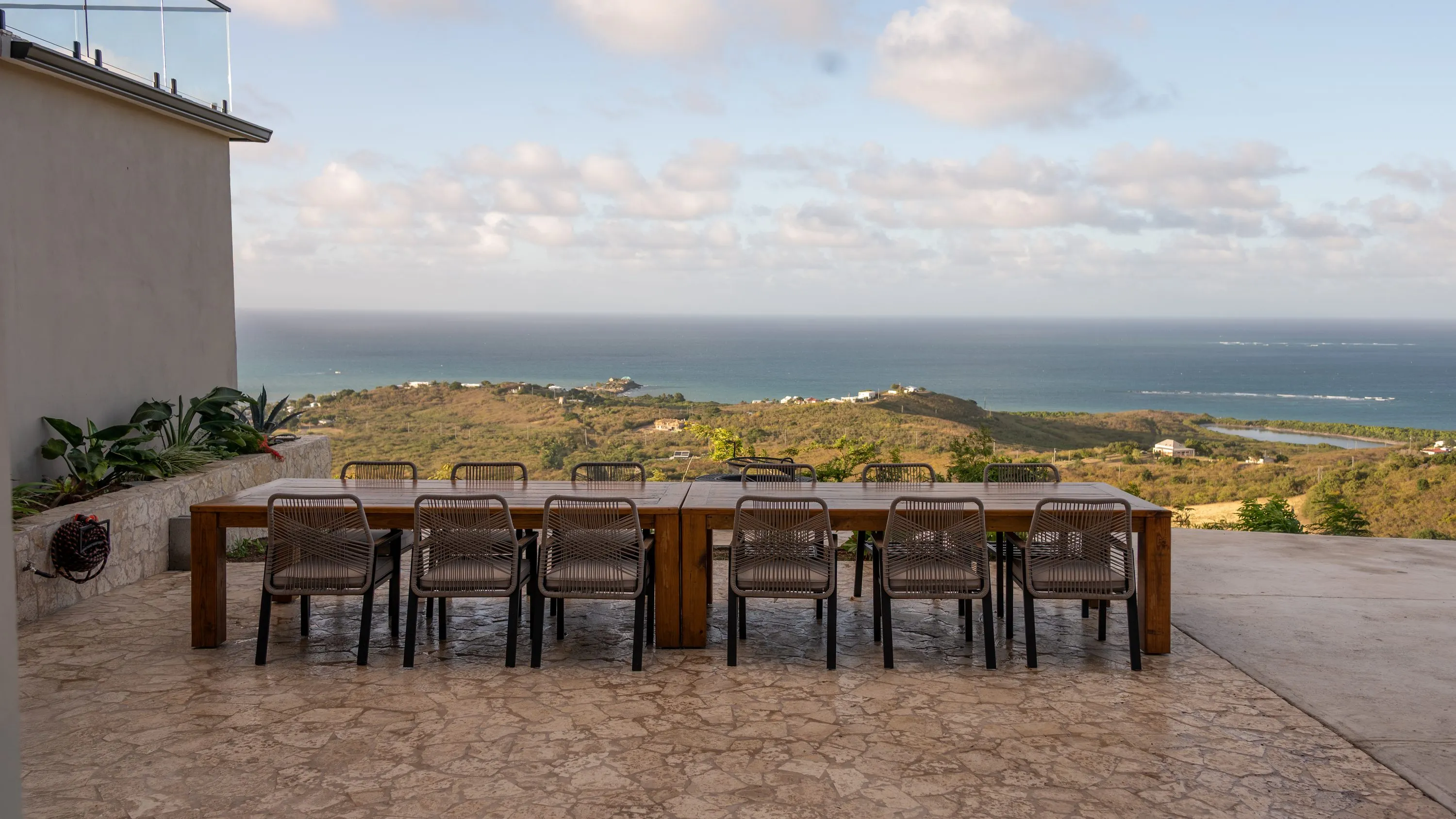 Long wooden dining table seating twelve on the stone terrace at golden hour, the north-east coast of St. Croix and the Caribbean Sea stretching behind it