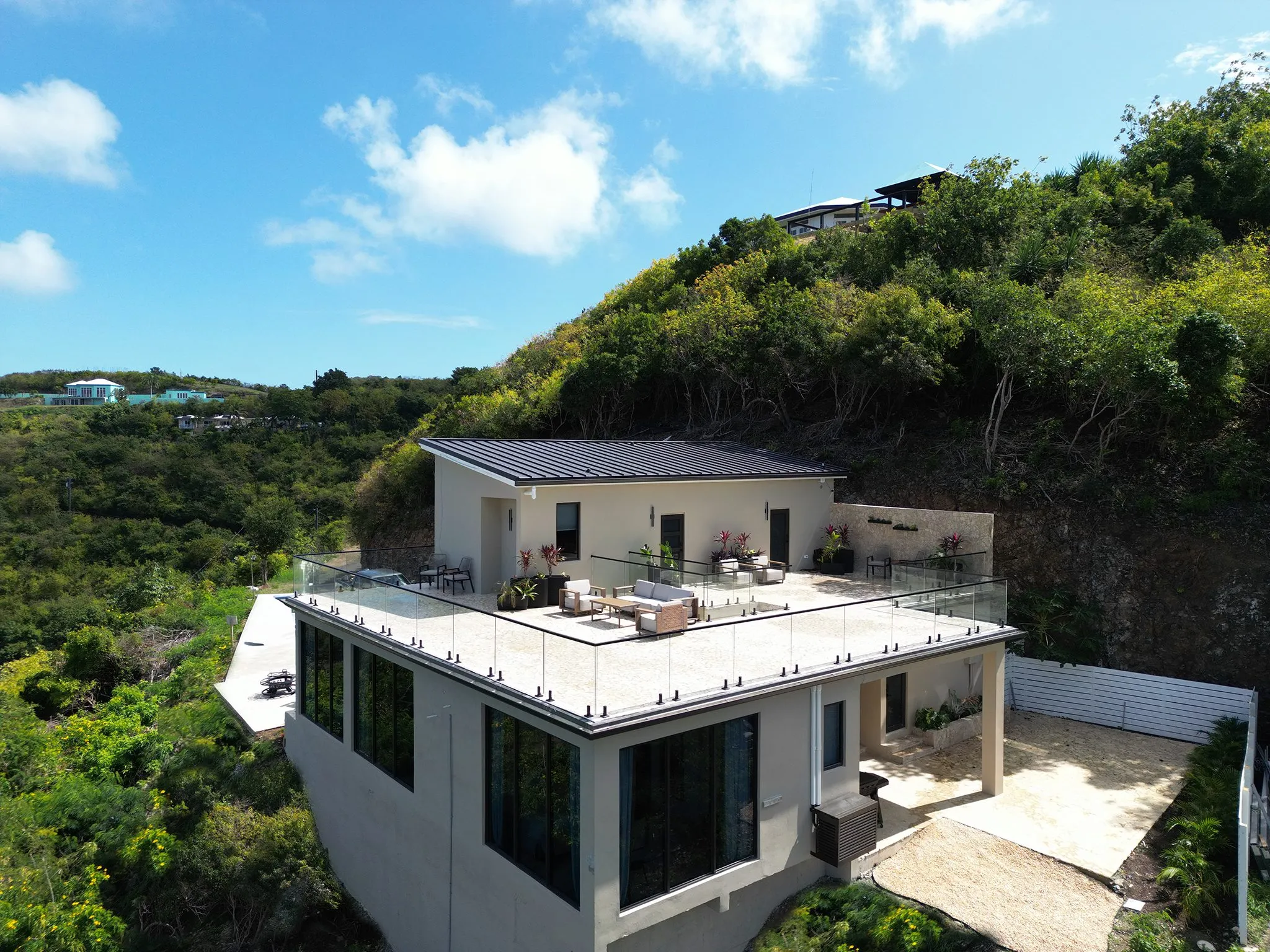 Aerial view of Crown of Caribbean — a white villa with a dark metal roof and glass-railed rooftop terrace, set into the green hillside on the east end of St. Croix